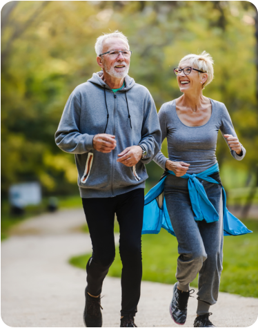 Photo of a senior woman sitting on a bench next to a walking trail in a park on a sunny day. She is holding her knee with a pained expression on her face.