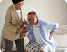 Photo of a senior woman sitting on a bench next to a walking trail in a park on a sunny day. She is holding her knee with a pained expression on her face.