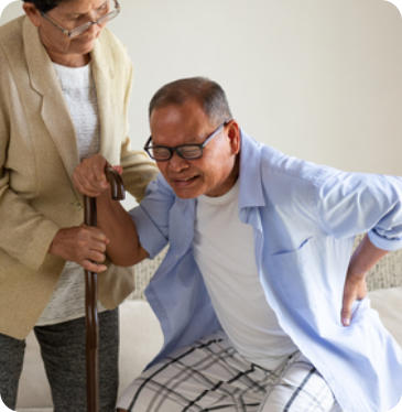 A senior woman is helping her husband stand up from a sitting position. He is grimacing in pain and holding his lower back.