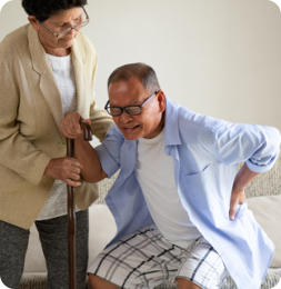 A senior woman is helping her husband stand up from a sitting position. He is grimacing in pain and holding his lower back.